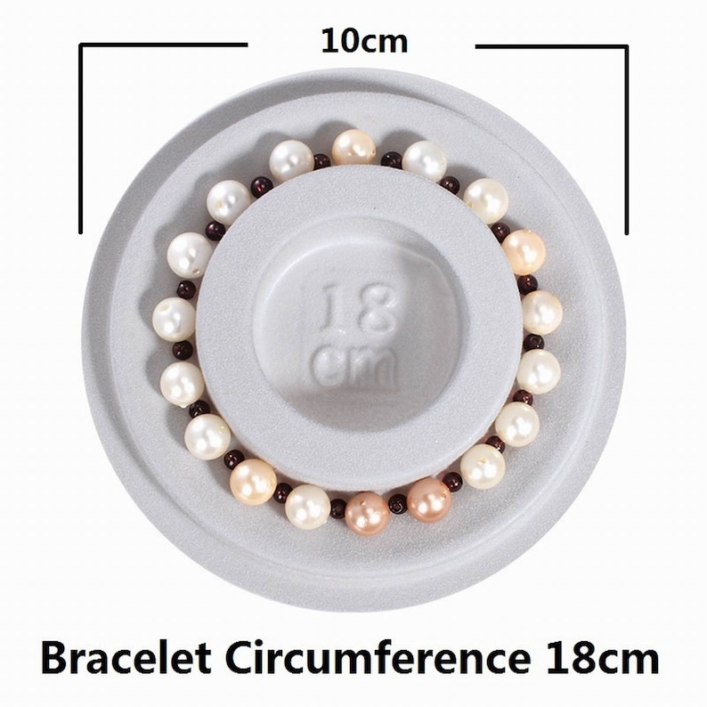May include: A white, gray, and brown pearl bracelet on a white display stand. The bracelet measures 18 cm in circumference. The stand is 10 cm in diameter.