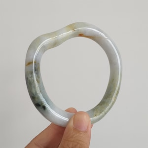 May include: A smooth, polished stone bangle bracelet with an irregular, wavy shape. The material is translucent and features a marbled pattern with shades of pale white, soft green, and small flecks of brown throughout the polished surface.