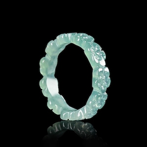 May include: A light green jadeite bangle bracelet. The bracelet is carved with a floral pattern. The jadeite has a translucent quality, and the bracelet is displayed against a black background with a reflection.