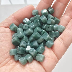 May include: A handful of jade beads in various shades of green. The beads are square-shaped with rounded edges and a hole drilled through each for stringing. The beads have a polished, glossy finish.
