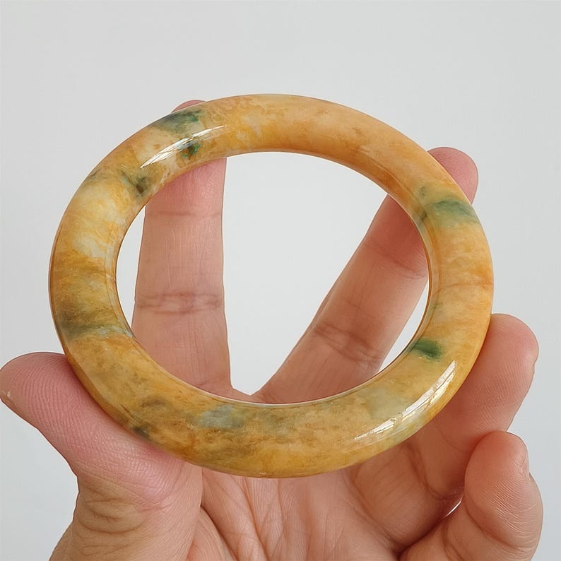 May include: A round, polished jade bangle bracelet. The bracelet features a blend of yellow, orange, and green hues. The jade is translucent, with visible veining and color variations. The bracelet is held in a hand against a plain background.