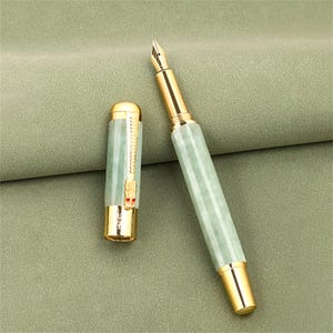 May include: A luxury fountain pen with a light green jade-like barrel and gold-colored accents. The cap is off, revealing the gold nib. The pen has a detailed design and is set against a green background.
