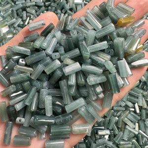 May include: A close-up shot displays a handful of small, cylindrical beads. The beads are a translucent, pale green color with darker green inclusions. They appear to be made of a stone-like material and are uniform in shape, with a hole running through the center.