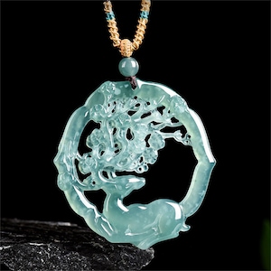 May include: A jade pendant necklace featuring a deer within a circular frame, carved with floral and tree branch details. The pendant is a light green color and is suspended from a gold-colored rope necklace.