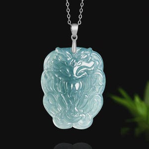May include: A light blue jade pendant carved in the shape of a lion's head, suspended from a silver chain. The pendant has intricate details and a translucent appearance. The background is dark, highlighting the pendant's color and design.