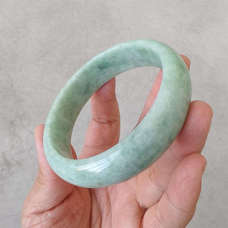 May include: A pale green jade bangle. The smooth, polished bracelet is a soft, light green colour. The jade is a solid, opaque stone. The bracelet is round and appears to be a standard size.