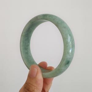 May include: A jade bangle bracelet in shades of green, held by a hand. The smooth, polished surface of the bracelet reflects light, showcasing its natural variations. The bangle's circular shape is clearly visible against a plain background.