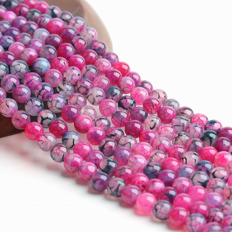 May include: Close-up of a collection of round beads in varying shades of pink and purple. The beads have a cracked, web-like pattern. The beads are strung together, suitable for jewellery making or other crafts.