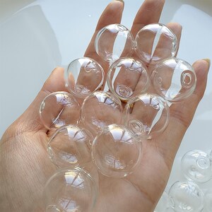 Op de afbeelding: Een hand vol heldere glazen bollen, elk met een klein gaatje. De bollen zijn rond en transparant en reflecteren licht. De afbeelding suggereert decoratieve elementen of knutselbenodigdheden. De bollen zijn waarschijnlijk in centimeters gemeten.