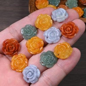 Op de afbeelding: Assortiment van jade rozenbloem bedels in verschillende kleuren, waaronder groen, rood, oranje en geel. De bedels zijn gegraveerd in een gedetailleerde rozenvorm en hebben een gladde, gepolijste afwerking.