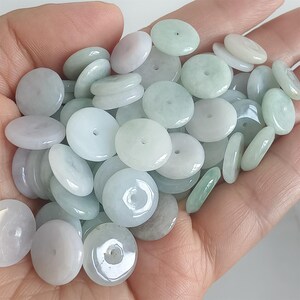 Könnte beinhalten: Eine Sammlung von Jadeperlen in verschiedenen Grün-, Weiß- und Lavendeltönen. Die Perlen sind rund und flach mit einem Loch in der Mitte und werden in einer Hand gehalten. Die Perlen haben unterschiedliche Größen und Formen und eine glatte, polierte Oberfläche.