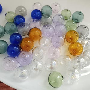 May include: A collection of spherical glass beads in various translucent colors including blue, amber, green, purple, and clear. Each bead features a small circular hole for threading, with smooth, reflective surfaces scattered across the frame.