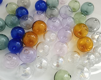 May include: A collection of spherical glass beads in various translucent colors including blue, amber, green, purple, and clear. Each bead features a small circular hole for threading, with smooth, reflective surfaces scattered across the frame.