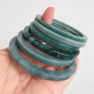 May include: A stack of four smooth, circular jade bangles in varying shades of deep forest green and muted sage. The bracelets feature polished surfaces with subtle natural variations in colour, ranging from solid opaque tones to mottled textures.