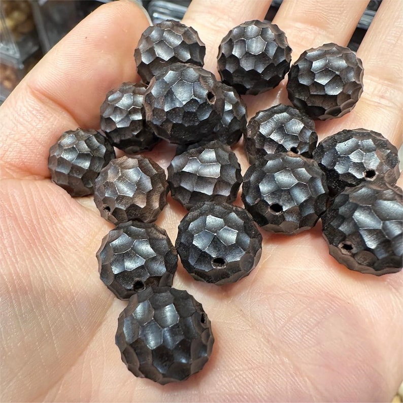 May include: A collection of dark brown, round, wooden beads with a textured surface. The beads are arranged in a pile on a hand.