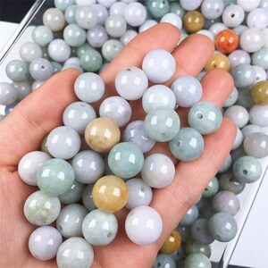 May include: A collection of round, smooth jade beads in various shades of green, white, and yellow. The beads are arranged in a hand, showcasing their size and texture.