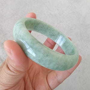 May include: A pale green jade bangle. The polished surface of the bracelet reflects light, highlighting its translucent nature. The jade has a soft, muted green hue, with slight tonal variations. This elegant bracelet is a classic piece of jewellery.