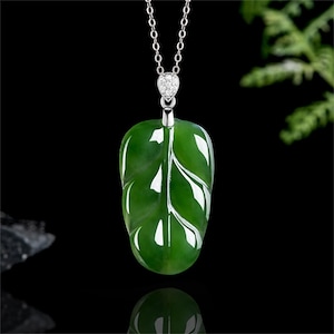 Puede incluir: Collar con colgante de hoja de jade verde y cadena de plata. El colgante en forma de hoja tiene vetas blancas y un acabado pulido. El collar se muestra sobre un fondo negro.