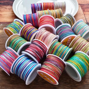 Può includere: Una collezione di rocchetti di filo colorato, ciascuno avvolto con più fili di colori vivaci. I rocchetti sono disposti su una superficie di legno, alcuni appoggiati su un piatto bianco. I fili sembrano essere per l'artigianato o la creazione di gioielli.