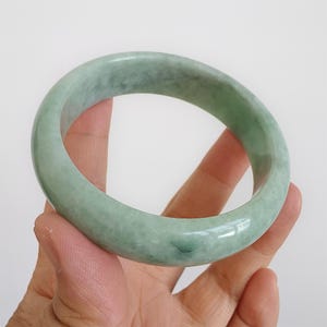 May include: A pale green jade bangle bracelet. The polished surface of the bracelet reflects light, highlighting its natural colour variations. The jade has a soft, muted green hue, with subtle veining. This classic piece of jewellery is a timeless accessory.