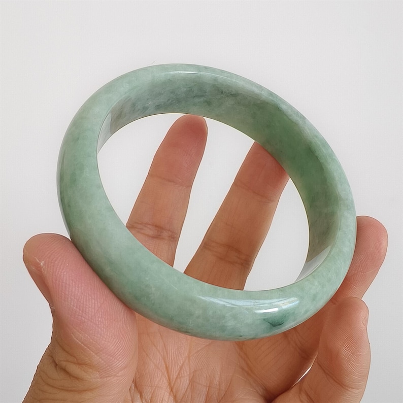 May include: A pale green jade bangle. The polished surface of the bracelet reflects light, highlighting its natural colour variations. The bangle is circular, designed to be worn on the wrist. A classic piece of jewellery.