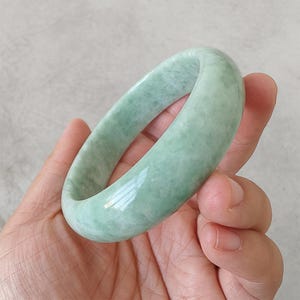 May include: A pale green jade bangle. The polished surface of the bracelet reflects light, highlighting its natural colour variations. The jade is a light green hue, with subtle marbling. A classic piece of jewellery, approximately 6.5 cm in diameter.
