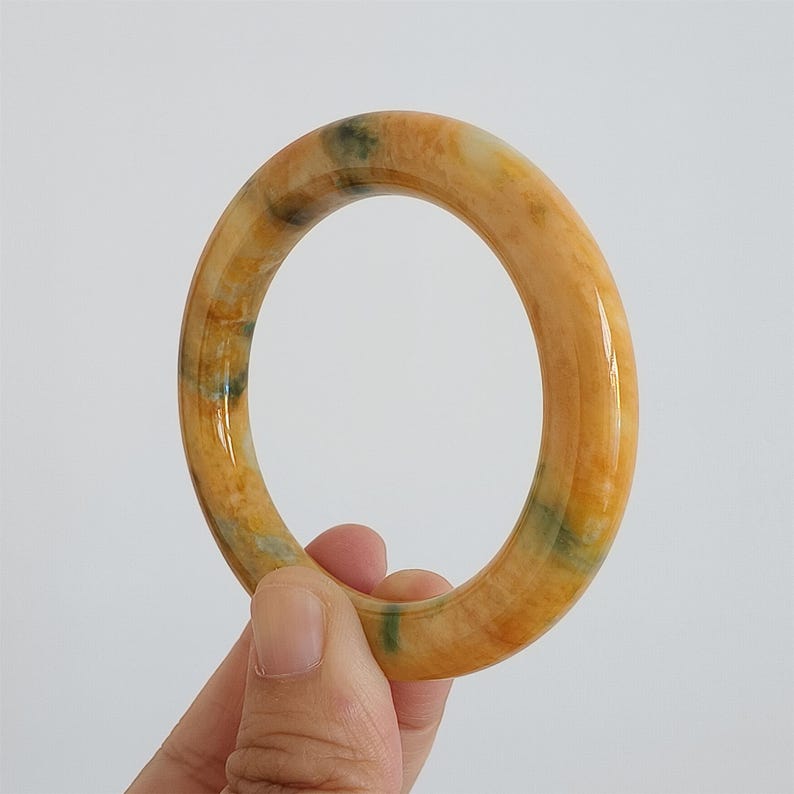 May include: A close-up of a jade bangle bracelet. The bracelet is a warm yellow-orange color with streaks of green and white. The smooth, polished surface reflects light, highlighting the natural variations in the stone. The bangle is held by a hand.