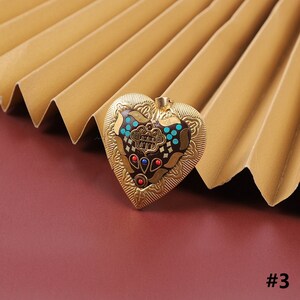 Puede incluir: Un colgante de coraz&oacute;n de oro con un dise&ntilde;o intrincado con esmalte marr&oacute;n, azul y rojo. El colgante tiene un patr&oacute;n floral detallado y una peque&ntilde;a pieza central adornada.
