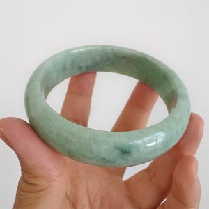 May include: A pale green jade bangle. The polished surface of the bracelet reflects light, highlighting its natural colour variations. The jade is a soft green with subtle darker green inclusions. The bangle's diameter is approximately 6.5 cm.