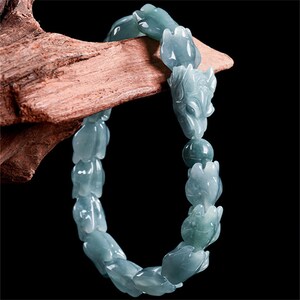 Puede incluir: Un brazalete de jade azul claro con cuentas talladas con forma de flores y animales. El brazalete est&aacute; ensartado y descansa sobre un trozo de madera, mostrando los intrincados detalles de la piedra preciosa.