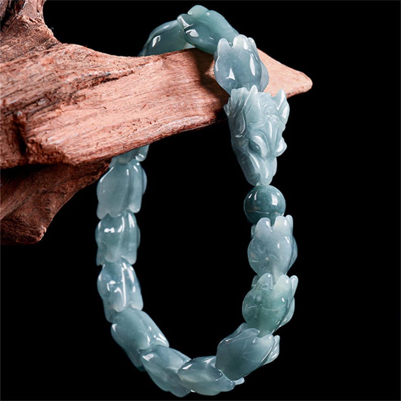 Puede incluir: Un brazalete de jade azul claro con cuentas talladas con forma de flores y animales. El brazalete est&aacute; ensartado y descansa sobre un trozo de madera, mostrando los intrincados detalles de la piedra preciosa.
