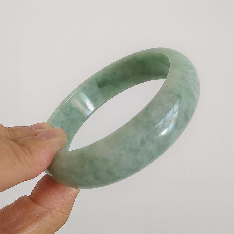 May include: A pale green jade bangle bracelet. The polished surface reflects light, highlighting the natural colour variations. The jade is a light green with subtle marbling.