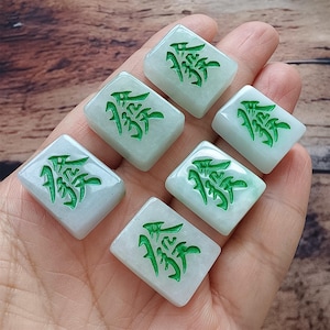 May include: Six rectangular jade tiles with a light green hue and a carved green symbol. The tiles are arranged in a hand, showcasing their polished surfaces and intricate design. The background is a wooden surface.