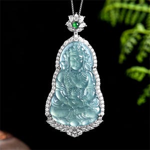 Könnte beinhalten: Eine Halskette mit Anhänger, die eine hellblaue Jade-Schnitzerei einer sitzenden Figur zeigt, eingefasst in Silber mit klaren Edelsteinen. Ein kleiner grüner Edelstein akzentuiert die Oberseite. Der Anhänger hängt an einer filigranen Silberkette.