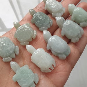 May include: A collection of small, carved jade turtle figurines in varying shades of green and white. The turtles are held in a hand, showcasing their detailed shell patterns and small heads and limbs. The jade has a translucent quality.