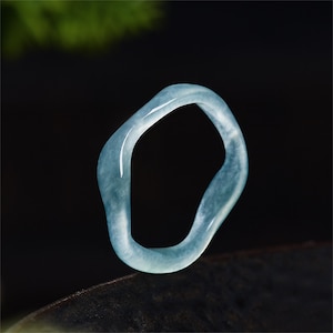 May include: A light blue, irregular-shaped ring. The ring is made of a translucent material, possibly jade or a similar stone. The ring's design is smooth and organic, with a polished finish. The ring is set against a dark background.