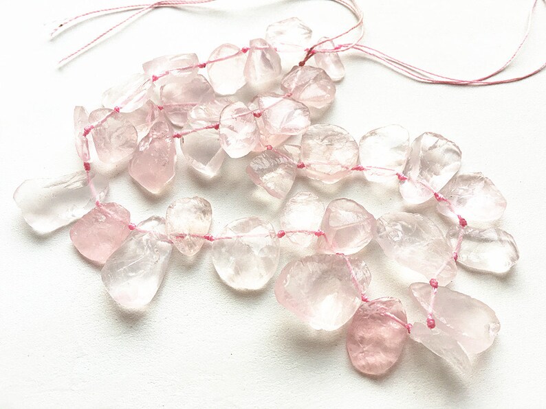 May include: A strand of pink rose quartz beads strung on pink thread. The beads are irregular in shape and have a translucent appearance.