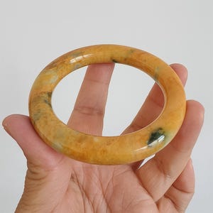 May include: A polished, round bangle bracelet in shades of yellow and orange with subtle green inclusions. The smooth, glossy surface reflects light, highlighting the natural variations in the stone. A classic jewelry piece.