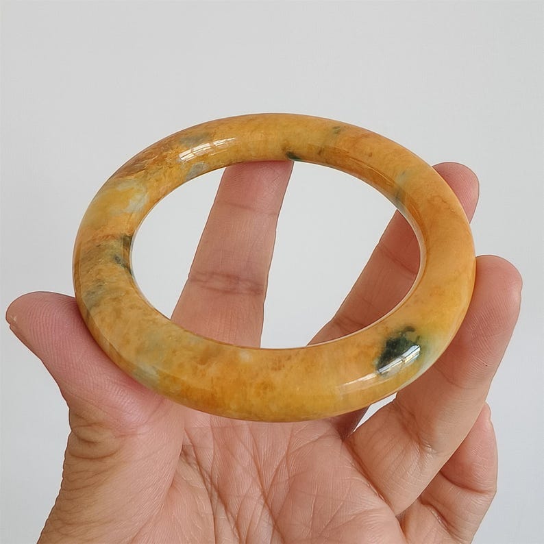 May include: A polished, round bangle bracelet in shades of yellow and orange with subtle green inclusions. The smooth, glossy surface reflects light, highlighting the natural variations in the stone. A classic jewelry piece.