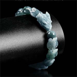 Puede incluir: Una pulsera de jade con cabezas de lobo talladas y cuentas en tonos azul-verdosos. La pulsera se muestra sobre un fondo negro, resaltando los intrincados detalles del tallado de la piedra preciosa. Una pieza de joyer&iacute;a &uacute;nica.