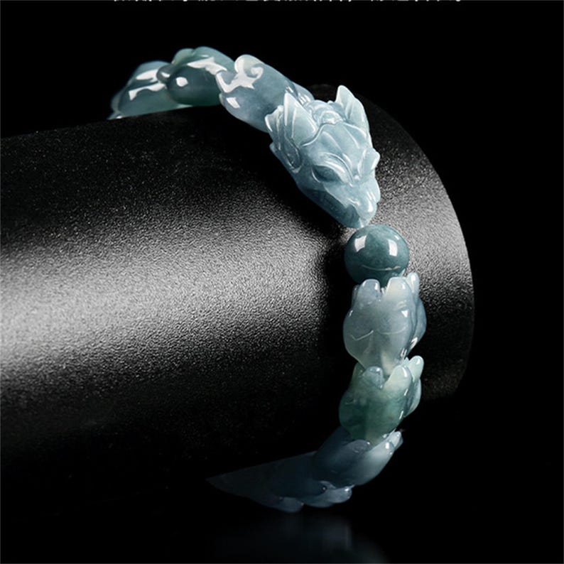 Puede incluir: Una pulsera de jade con cabezas de lobo talladas y cuentas en tonos azul-verdosos. La pulsera se muestra sobre un fondo negro, resaltando los intrincados detalles del tallado de la piedra preciosa. Una pieza de joyer&iacute;a &uacute;nica.