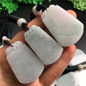 Könnte beinhalten: Drei Jade-Anhänger mit rechteckiger Form und abgerundeten Kanten werden gezeigt. Jeder Anhänger hat eine hellgrüne Farbe mit einem weißen Farbverlauf. Die Anhänger sind an schwarzen Kordeln mit weißen Perlen und grünen Akzenten befestigt.