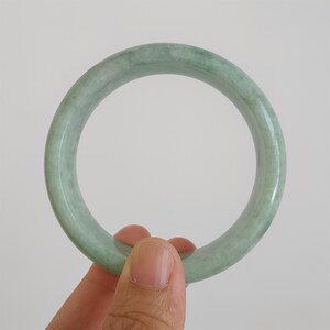 May include: A pale green jade bangle. The polished bracelet is held by a hand. The jade has a translucent quality with subtle colour variations. The bracelet is a circular shape.