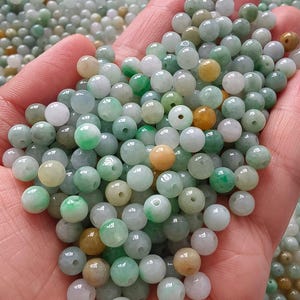 May include: A collection of small, round, polished beads in various shades of green, white, and light yellow. Each bead features a central hole for threading. The beads have a smooth, semi-translucent appearance with natural colour variations.