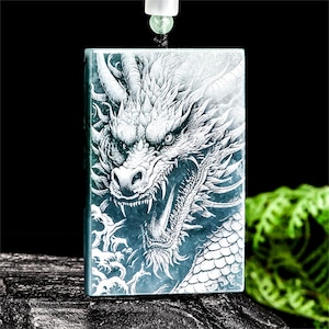 Puede incluir: Colgante rectangular con un detallado grabado de la cabeza de un dragón. El dragón es blanco sobre un fondo verde azulado, con escamas intrincadas y una expresión feroz. El colgante cuelga de un cordón negro con una cuenta verde.