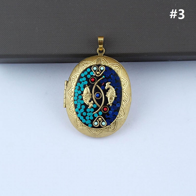 May include: A gold oval locket with a blue and turquoise mosaic design featuring two gold fish. The locket has a hinge on the side and a loop at the top for attaching a chain.