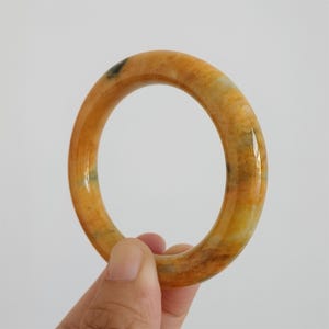 May include: A polished, circular bangle bracelet in shades of yellow, orange, and brown. The bracelet has a smooth, glossy finish and is held by a hand against a white background. The bangle is a piece of jewelry.