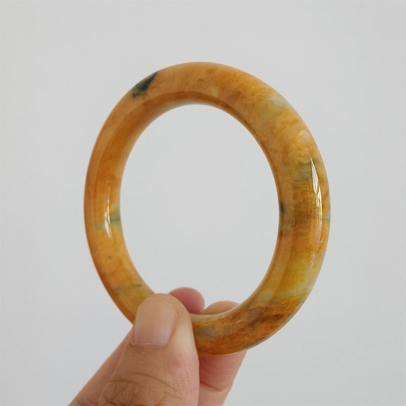 May include: A polished, circular bangle bracelet in shades of yellow, orange, and brown. The bracelet has a smooth, glossy finish and is held by a hand against a white background. The bangle is a piece of jewelry.