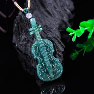 May include: A teal-colored violin pendant carved from jade, suspended on a cord. The pendant features intricate details, including the instrument's strings and body design. The background is a dark, textured surface with a hint of green foliage.