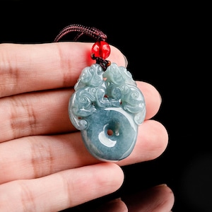 May include: A jade pendant necklace with a green jade pendant carved with two dragons. The pendant is attached to a brown cord with a red bead.